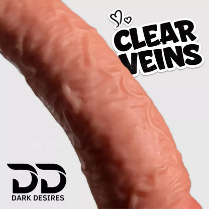 A close-up of a textured skin -beige curved object with printed vein patterns and realistic textured dildo with a black harness-style strap, 10-inch curved Extra Thick strap-on dildo with suction cup and adjustable harness for G-spot, vaginal and anal play Black faux harness leather belt Sex Toys for Women in Mumbai India by DarkDesires
